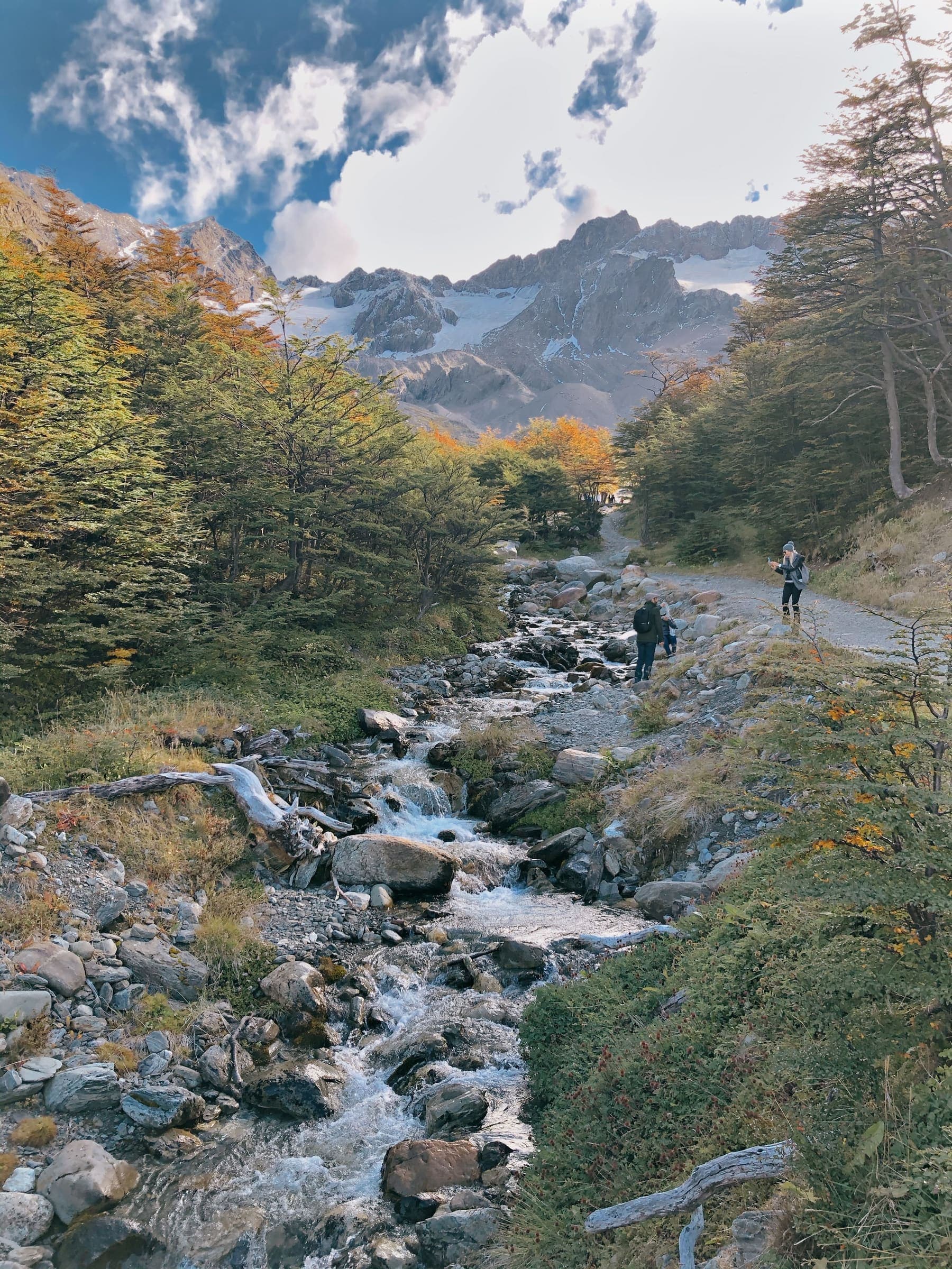 Hikers on a trail beside a stream under snow peaks